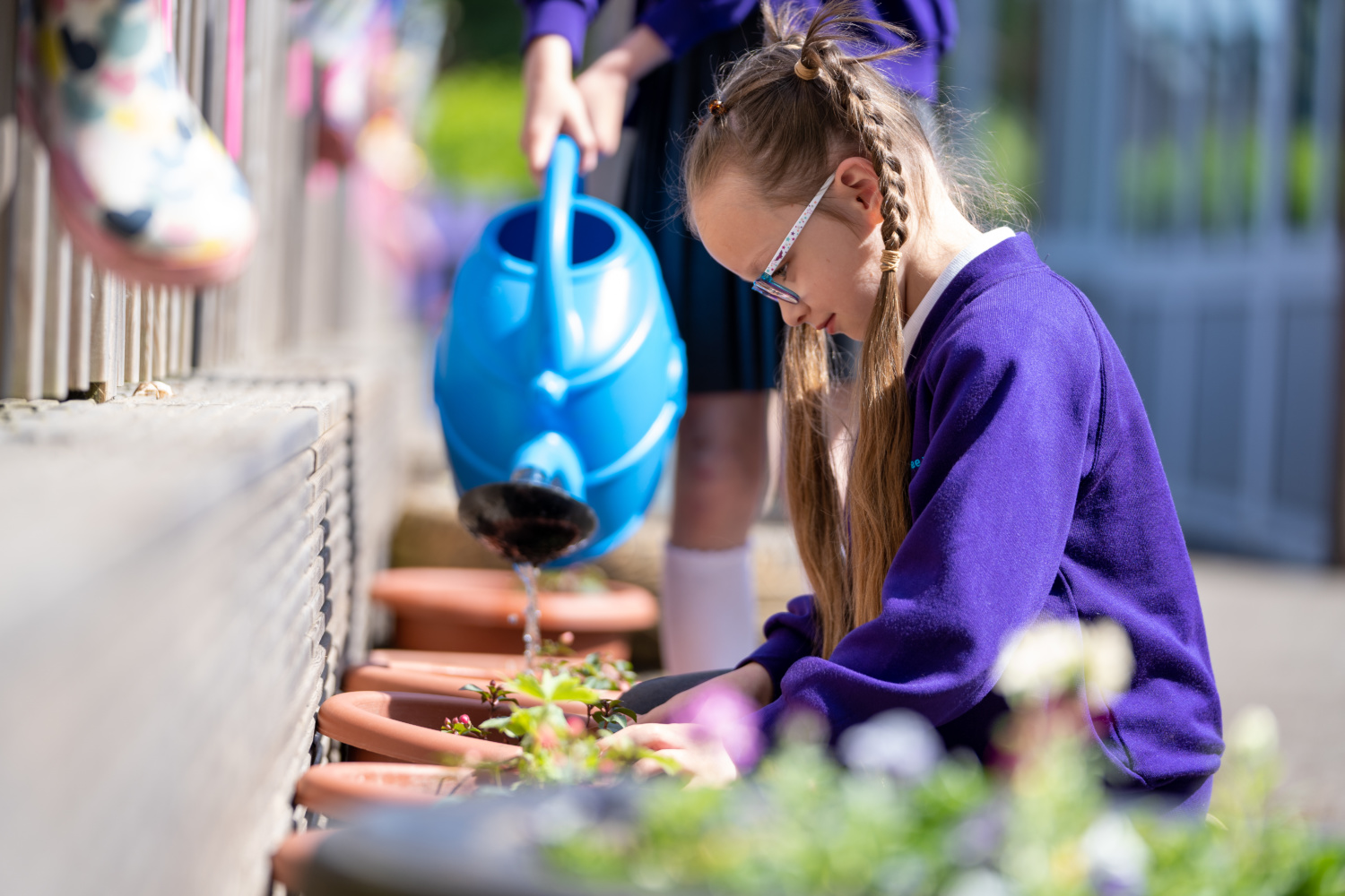 A young girl is seen dressed in her academy uniform, gardening in the school outdoor play area, whilst a friend is pictured watering the plant pots nearby.